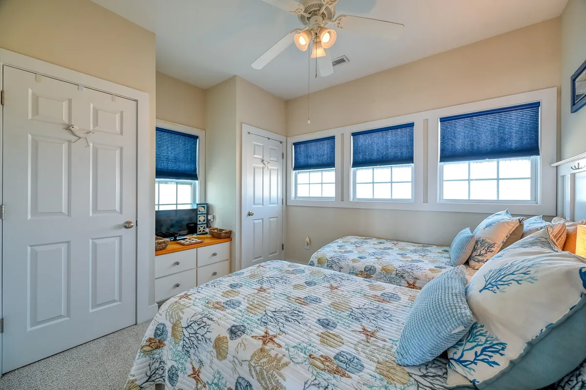 This image depicts a tranquil guest bedroom with twin beds featuring coral-themed bedding, blue window shades, and a small desk with a television. The setup reflects practical built-in storage and flexible sleeping arrangements ideal for families or friends.