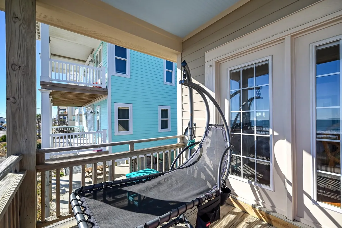 A hammock strung on a wooden deck outside a blue house with a view of the surrounding landscape. The calm setup evokes a sense of solitude and relaxation, ideal for reading or meditating while immersed in nature.