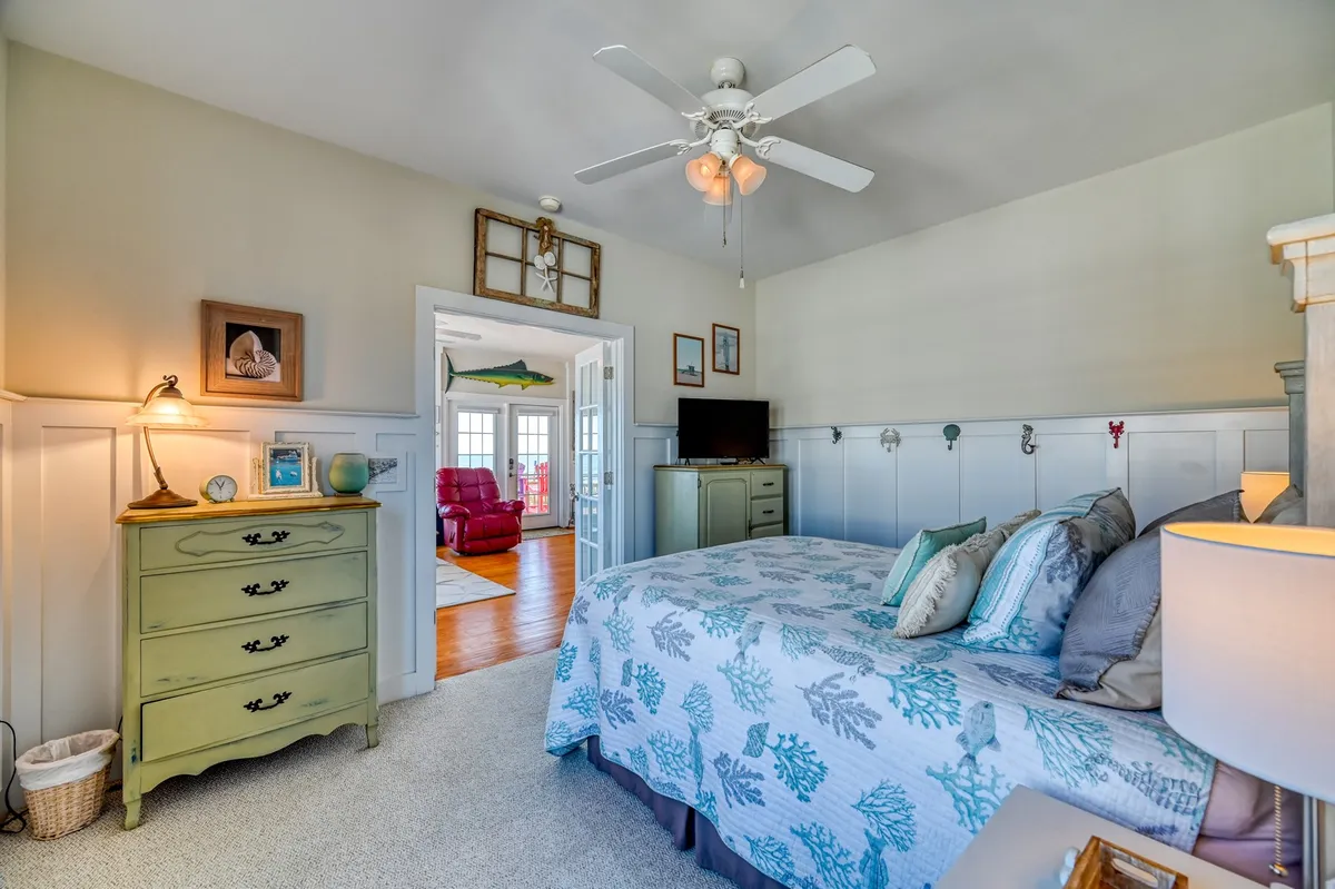 This image showcases a cozy bedroom with a bed adorned in decorative bedding, a vintage dresser, and a doorway that hints at connected living spaces. Natural light fills the room, highlighting its warm, welcoming ambiance and classic cottage character.
