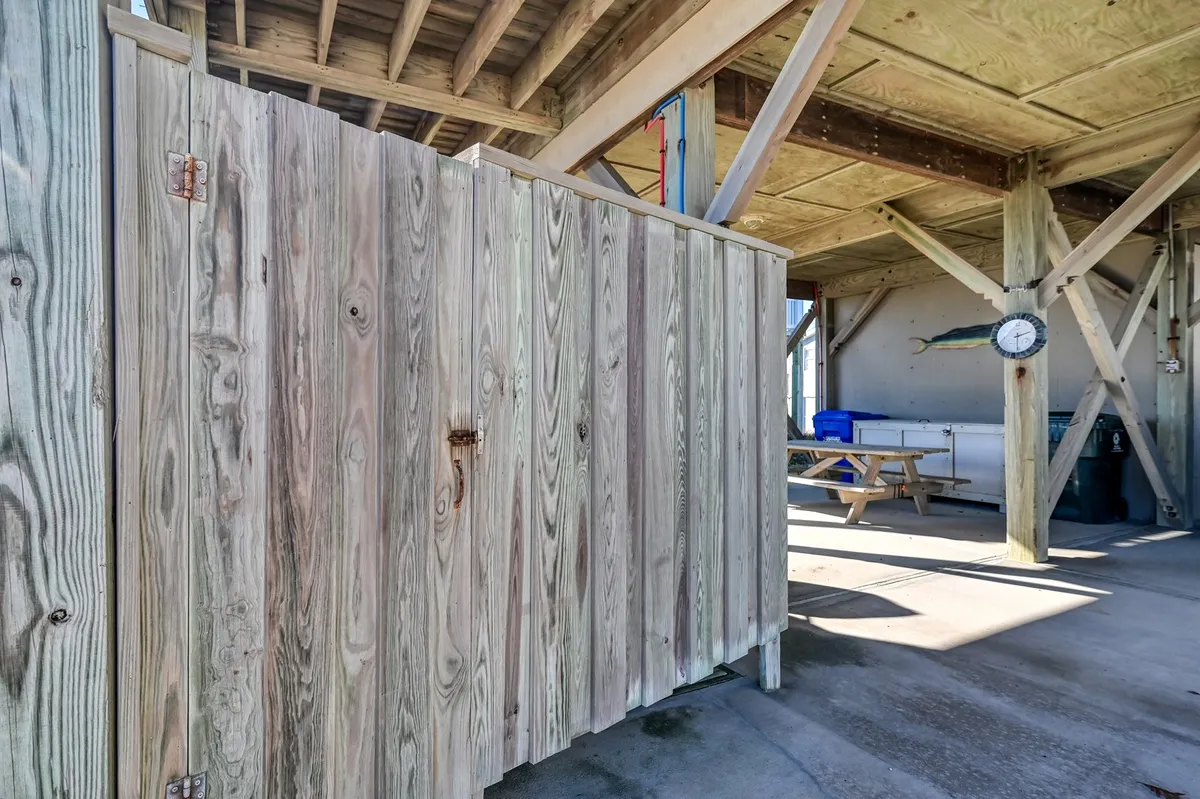 An exterior view of a wooden outdoor shower enclosure and a shaded picnic table, with subtle fishing décor and a sense of relaxed coastal living. The clock and picnic table reinforce the practical outdoor amenities.
