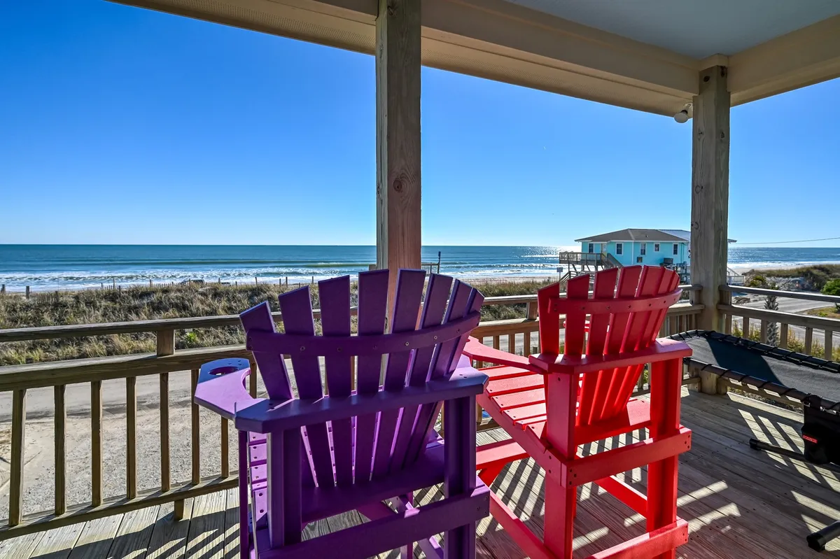 Two vibrant adirondack chairs overlook the ocean from a porch, with a sweeping view of the waves and sky. The setting is perfect for reading, watching the sunrise, or simply soaking in the peaceful atmosphere.