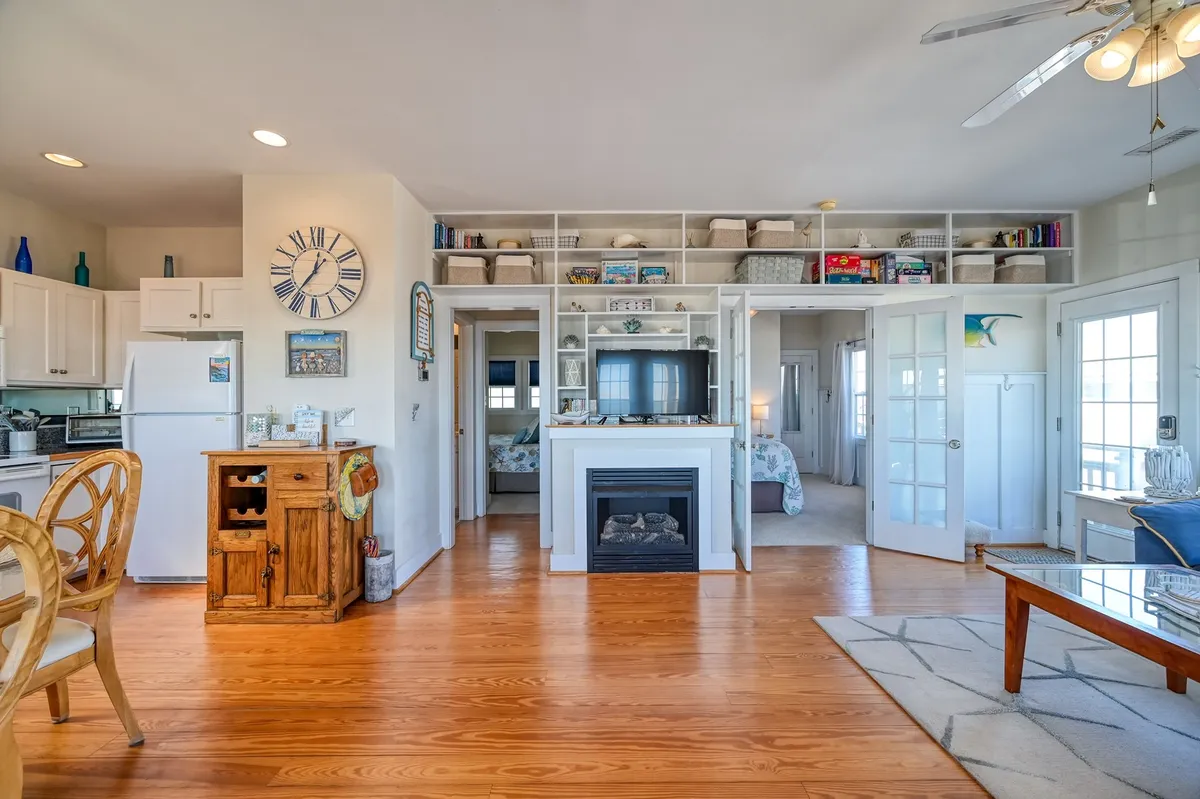 A warmly lit living area with a central electric fireplace, open layout, dining area, and shelves with decorative coastal touches. The space looks cozy and inviting, perfect for relaxing by the fire or gathering for meals during cooler months.