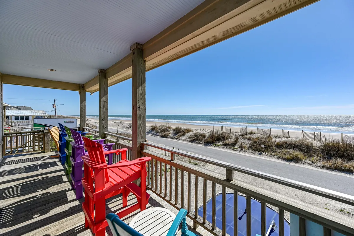 A vibrant exterior porch scene with colorful Adirondack chairs overlooking sand dunes, the ocean, and a blue sky. The setting feels cheerful and perfect for outdoor relaxation in any season.
