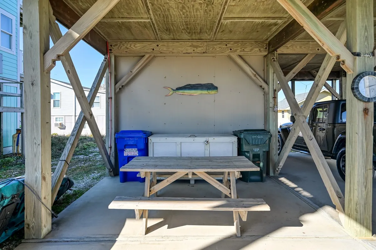 An exterior scene highlighting a picnic table, blue recycling bin, white storage unit, and a playful fish mural. This setting is ideal for group activities like outdoor dining, barbecues, or casual games, with an easygoing, beachy vibe.