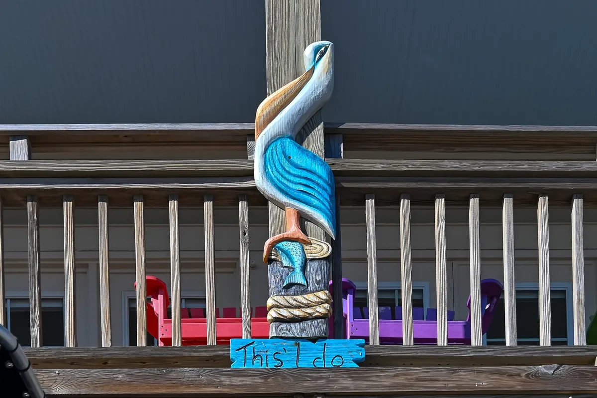 This exterior image shows a decorative wooden pelican carving, with colorful Adirondack chairs arranged for socializing, and the words 'This I do' creating a whimsical, welcoming touch. The open-air setting overlooks a likely beach or deck area.