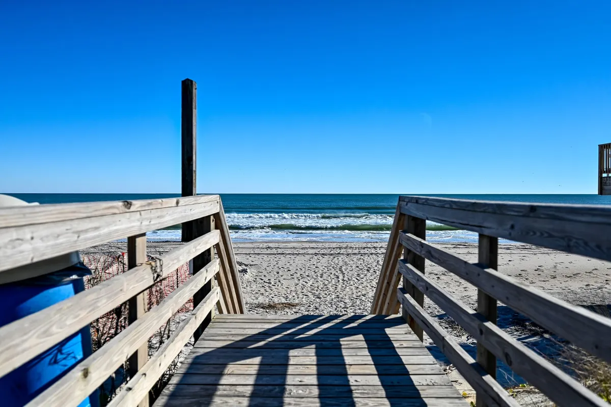 An exterior shot of a wooden walkway leading directly to the ocean, bordered by blue sky and dunes. The image evokes the unique luxury of having a private crossover moments from the sand.