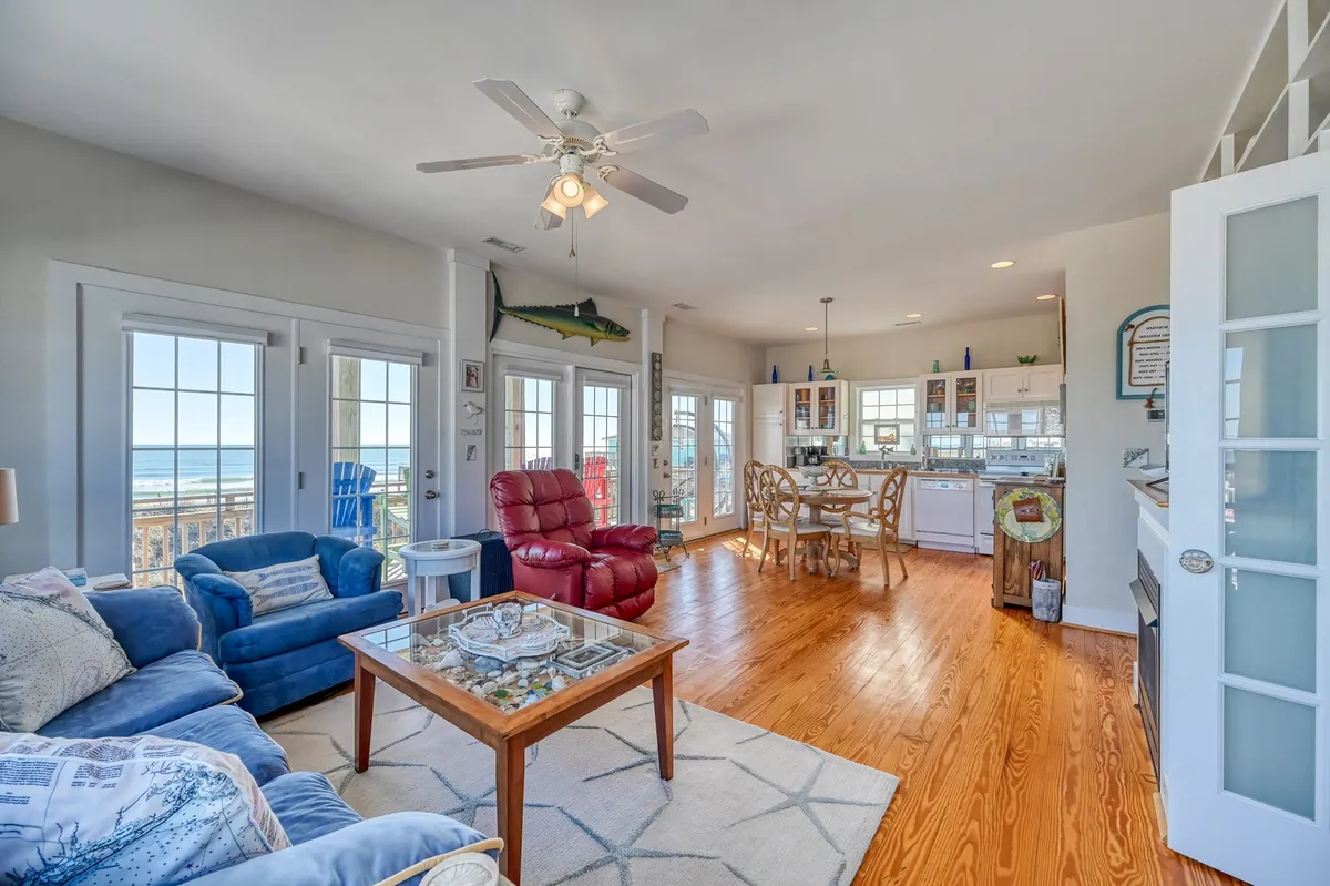 A cozy, open-concept interior with large windows that offer ocean views, a comfortable blue sofa, a red recliner, a dining table, and a kitchen area. The space is sunlit and designed for both relaxing and entertaining, with ample room for family gatherings or casual dining.