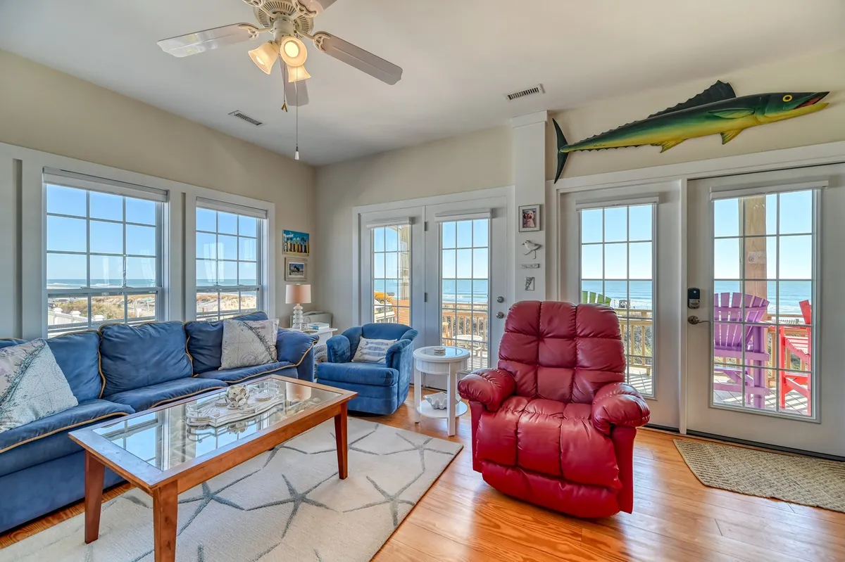 A sunlit living room with plush blue sofas, a bold red recliner, and playful fish décor, all facing wide open windows framing endless Atlantic Ocean views just beyond the dunes.