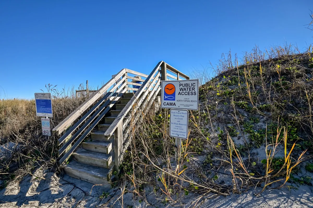 A wooden staircase leads over grassy dunes towards the sand, flanked by public access signs and surrounded by children playing. The relaxed setting and clear path to the beach highlight the convenience and safety of the location for families eager to reach the shoreline.