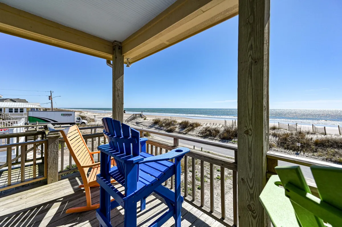 A lineup of colorful Adirondack chairs faces the wide stretch of white sandy beach and ocean. The clear, blue sky and gentle waves create a cheerful, inviting coastal atmosphere. The beach appears easily accessible, perfect for families looking to relax, socialize, or embark on seaside adventures just steps from their accommodation.