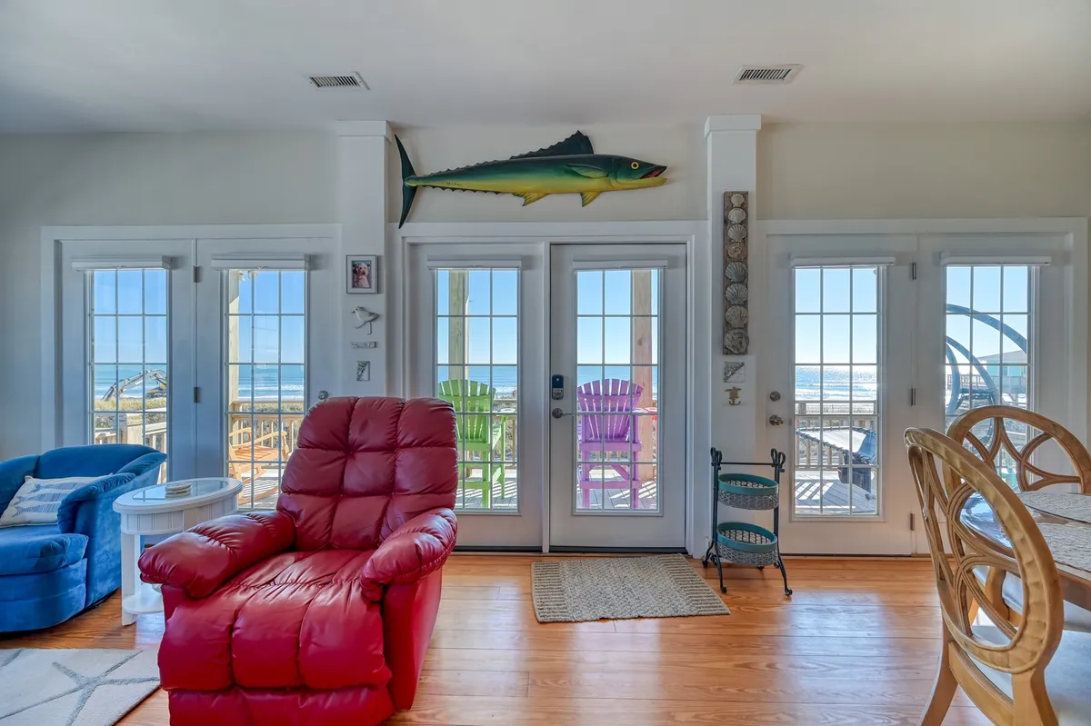 A cheerful interior scene featuring a red reclining chair, blue sofa, colorful Adirondack chairs, and large glass doors opening onto a deck with an ocean view. Decorative elements evoke a coastal feel.