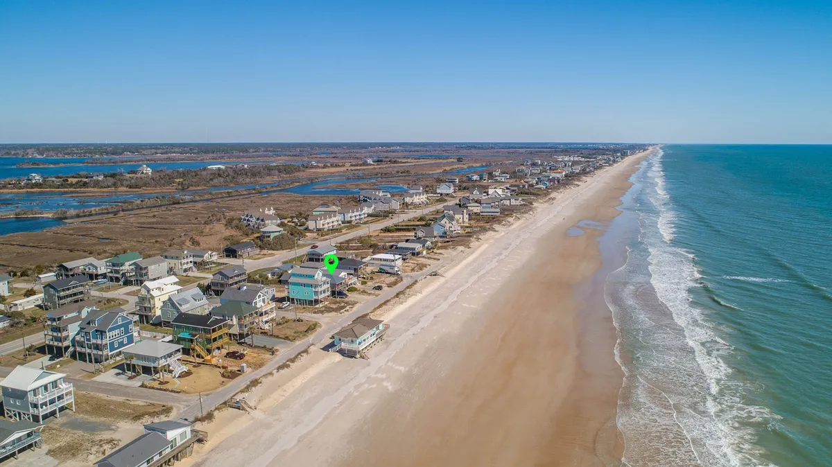 A broad view of beachfront property with rolling ocean waves, residential homes, and a classic coastal landscape. The scene features expansive sand, gentle surf, and ample space for activities like sandcastle building and shell hunting—the ideal setup for memorable family beach days.